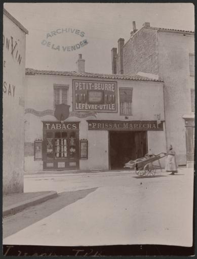Un magasin de tabacs et l'atelier "Prissac Maréchal" au 69 rue de La Roche, avec une publicité pour le petit-beurre Lefèvre-Utile sur la façade. Une vendeuse avec une charrette à bras passe devant l'atelier.